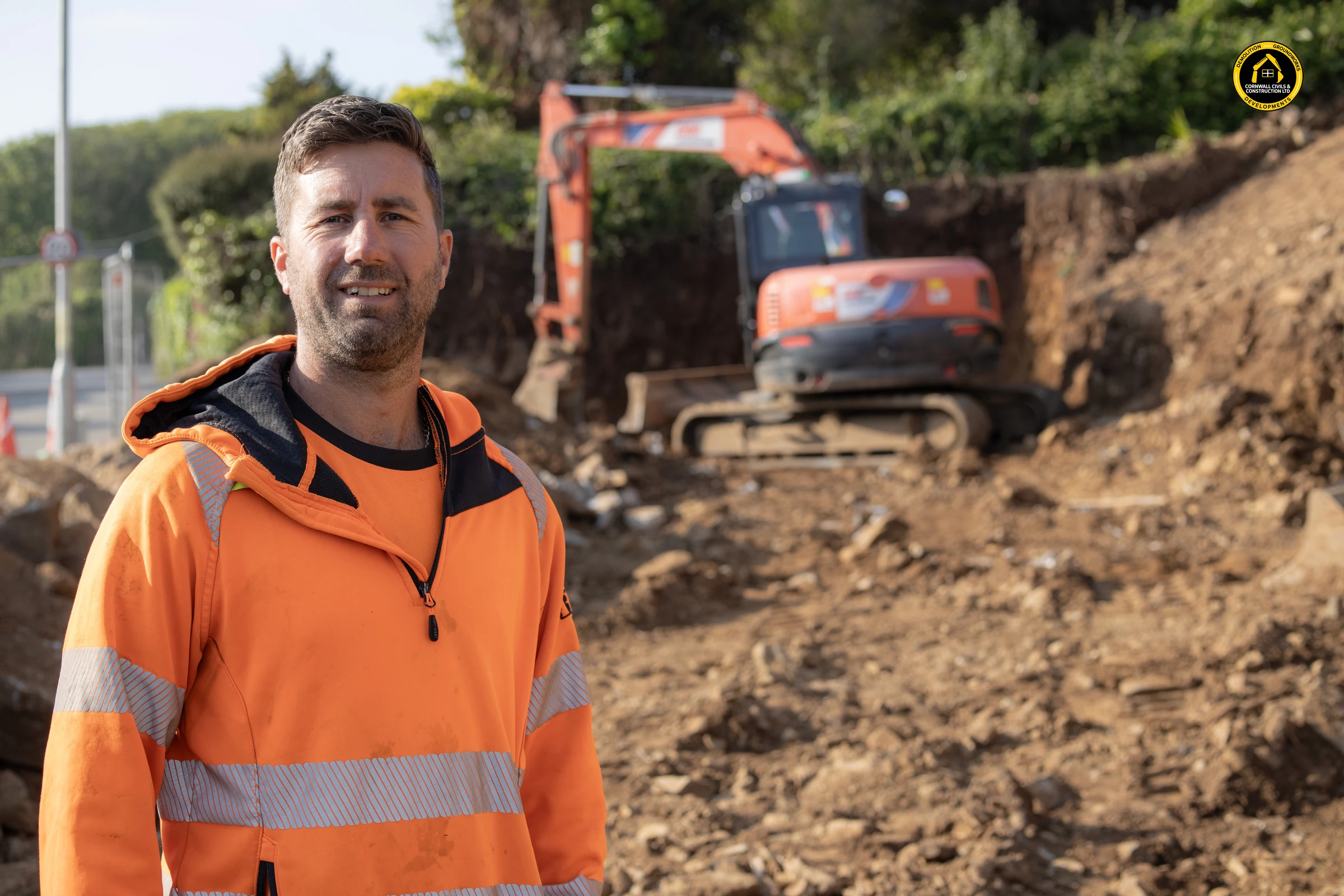 Matt Richards of Cornwall Civils & Construction on site in hi-vis, with his excavator working the dig behind him
