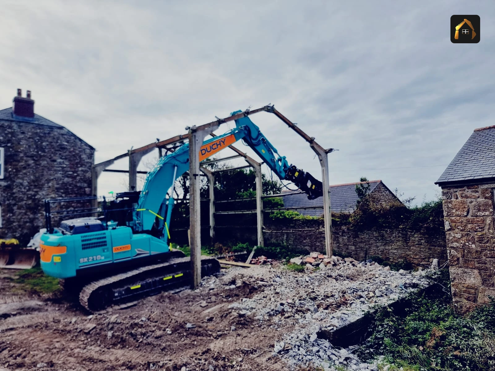 Tracked excavator demolishing a steel framed barn beside a granite cottage in Pendeen