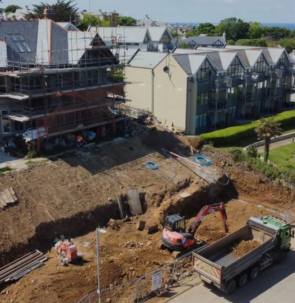 Excavator loading a tipper on the St Ives groundworks site beside a scaffolded building, with the sea behind